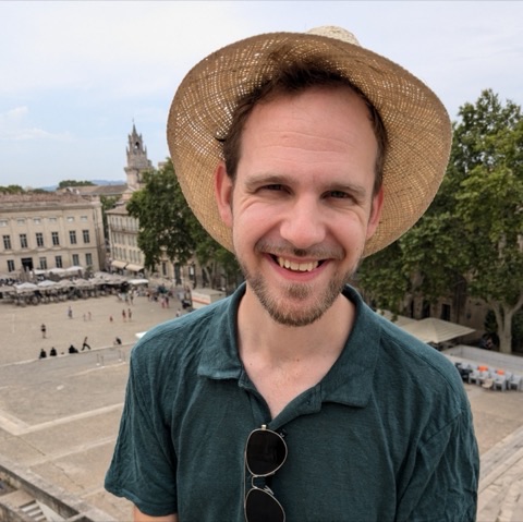Maarten smiling in a straw hat, a sunlit town square behind him